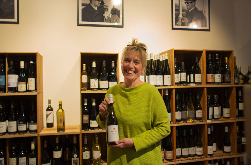 Eine lachende Frau präsentiert eine Weinflasche vor einem Weinregal